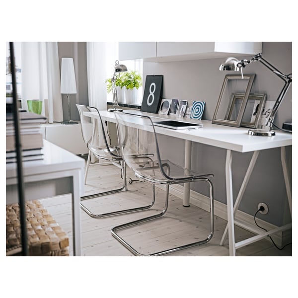 Modern home office with white desk, clear chairs, and sleek lamp. Light grey walls, wooden floors, and a cosy blanket.