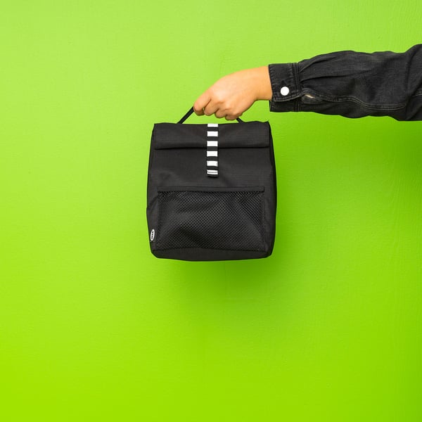 Person holding black rectangular lunch bag with mesh pocket against green wall.