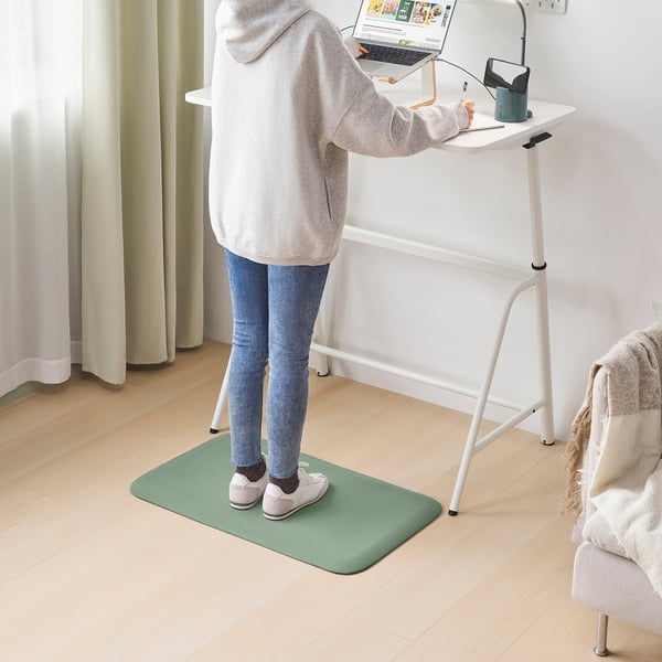 A person using a FREIVID green recycled standing mat in an office setup with a nearby chair and blanket.