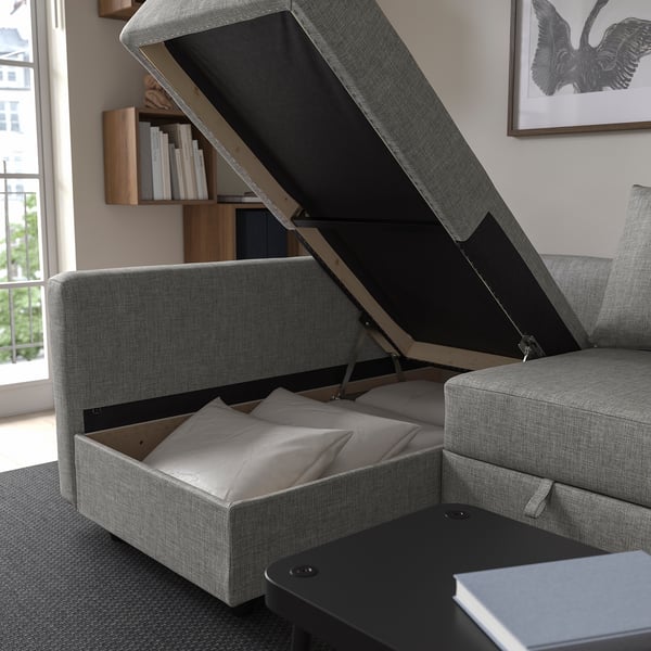 A grey upholstered sofa with open storage below, holding folded bedding and pillows.