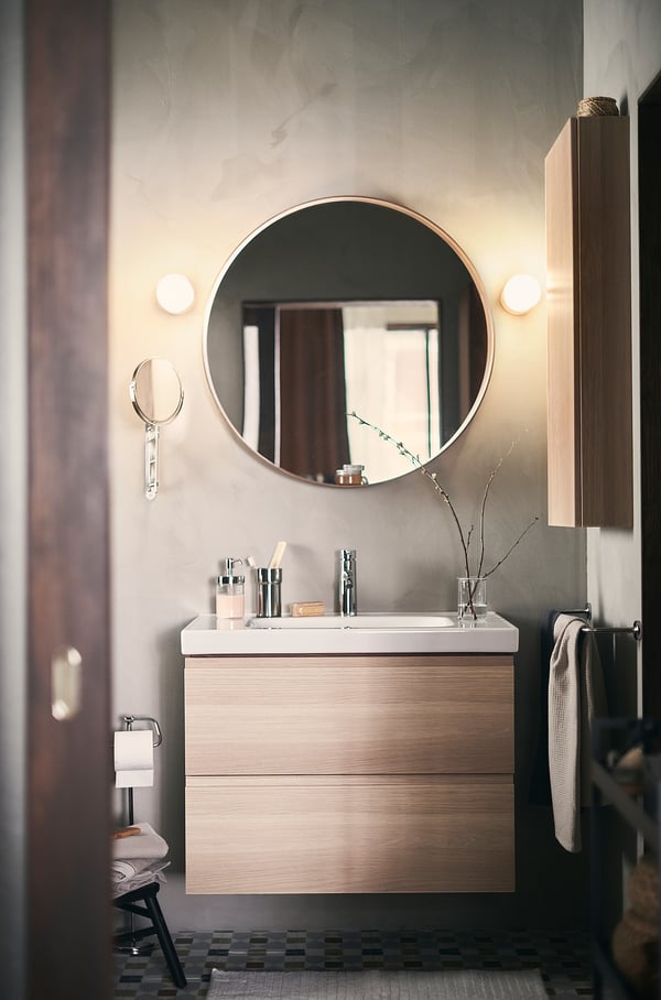 Modern bathroom with floating light wood vanity, round mirror, chrome fixtures, and black and white tiled floor.
