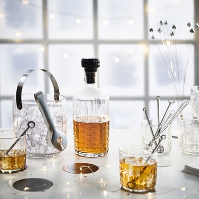 Cocktail setup: clear glass bottle with black cap, ice bucket, tongs, glasses, stirrers on white table.