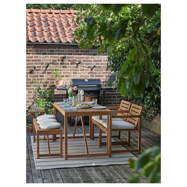 Outdoor dining setup with wooden table and four cushioned chairs on a patio. Brick wall and grill in background.