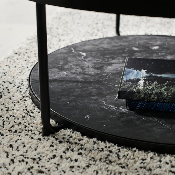 Black round side table with dark glass top, metal legs, and a book on it, on a spotted rug.