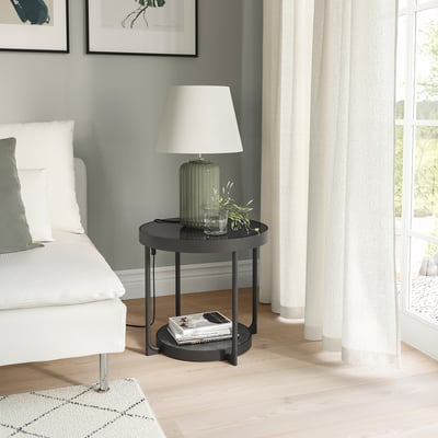 Modern black round side table with glass top next to white bed and large window.