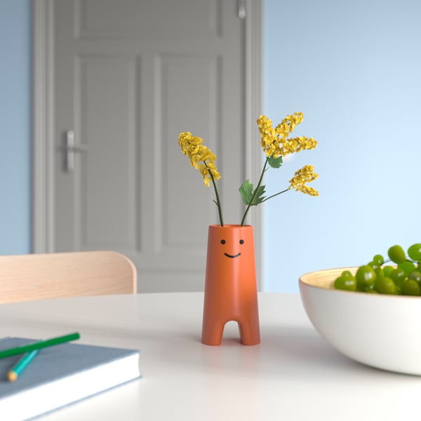 Orange FRÖVÄXT vase with smiling face, small, holds yellow flowers, on white table by bowl of grapes.