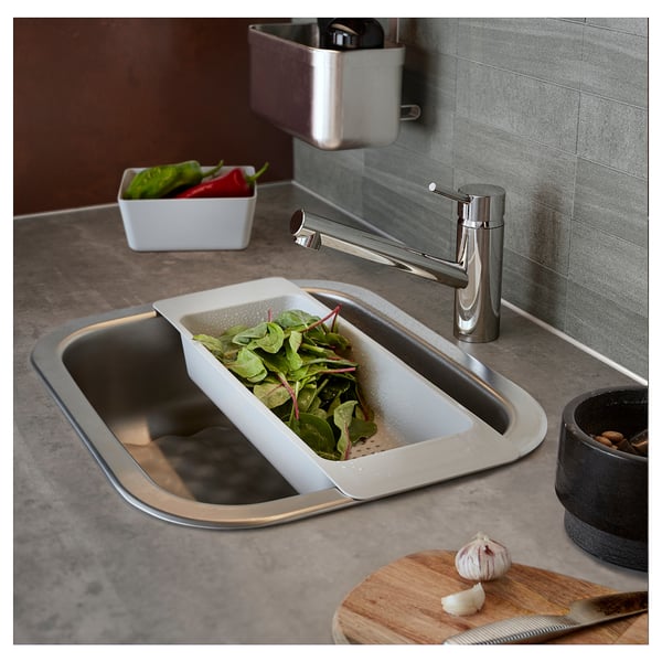 Modern kitchen sink with stainless steel faucet. White colander with green vegetables in sink. Wooden cutting board with knife and garlic nearby.