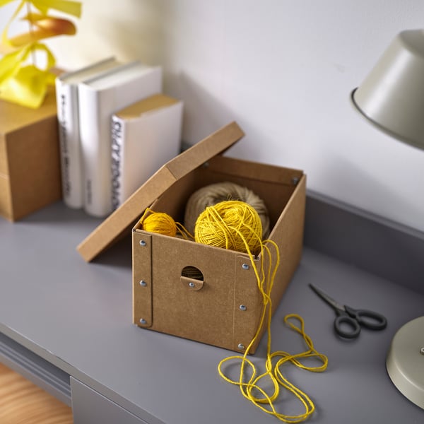 Small yellow yarn balls in brown cardboard box, on grey desk with books and scissors.