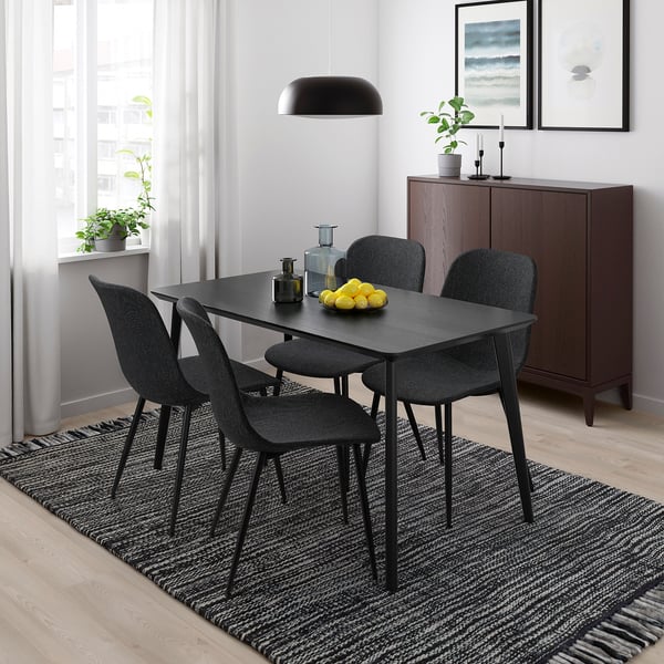 Modern dining setup with recycled GALLERUD chairs and table. Bowl of lemons, bottle, and vase on a striped rug. Dark cabinet and plants enhance decor.