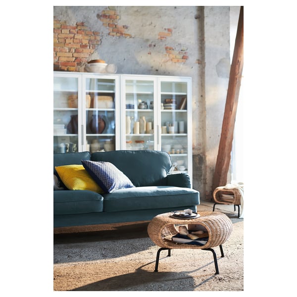 Blue GAMLEHULT footstool, woven rattan, round shape, rustic design, beside a green sofa, near a brick wall, under warm lighting.