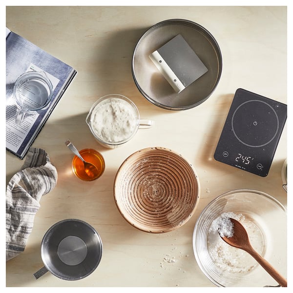 Kitchen with baking tools: scale, banneton, cups, honey, spoon, cloth, book, dough scraper.