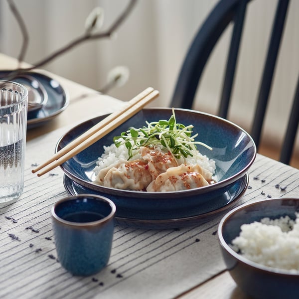 Blue ceramic bowl with dumplings, rice, and greens, wooden chopsticks, small cup, and glass on a striped table mat.