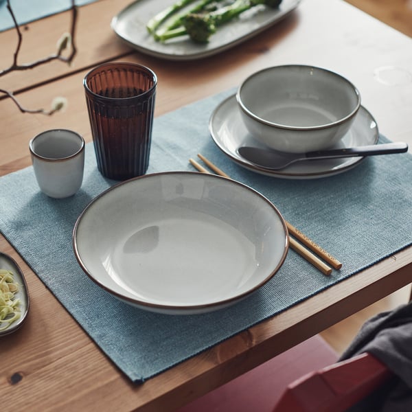 GLADELIG deep plate, sand-colored & stoneware, sits on a blue mat, with chopsticks, cup, and spoon nearby. Larger than surrounding items.