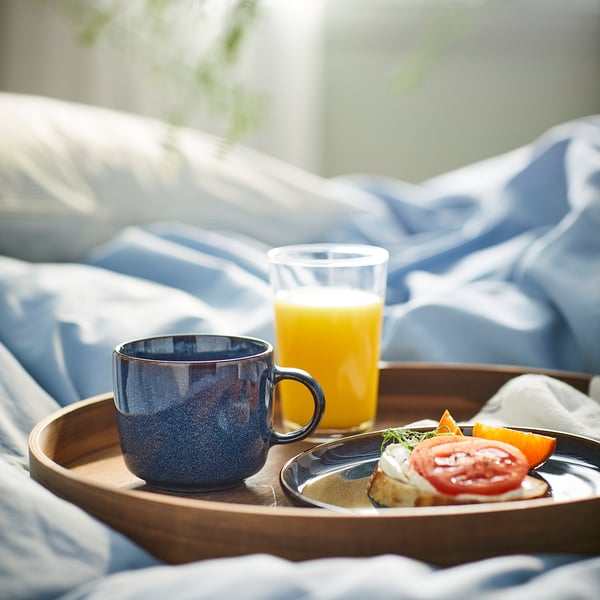 Blue stoneware mug, orange juice, toast on wooden tray against blue bedspread.