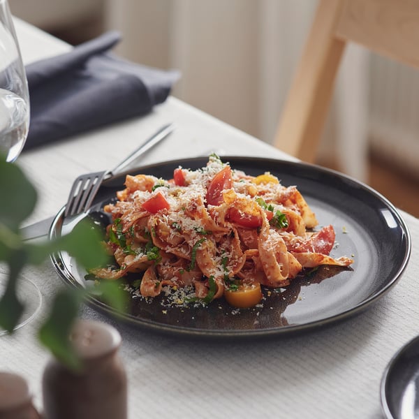 Plate of pasta with cherry tomatoes and grated cheese on a dark plate with fork, set on dining table.