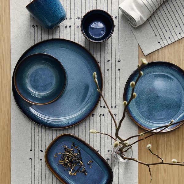 GLADELIG table setting: deep blue plates, bowl, and mug. Stoneware material. Centred on light, striped mat. Branched floral decoration.