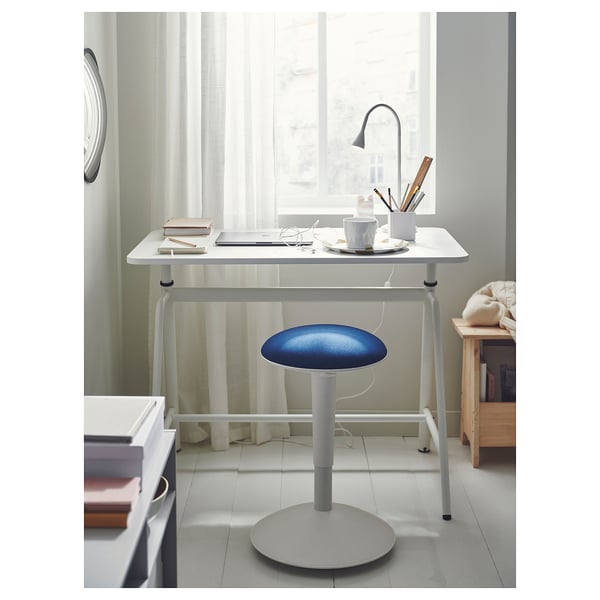 White sit/stand desk with blue stool, lamp, laptop, and coffee cup.