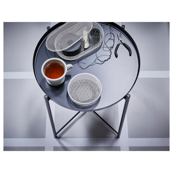 Small black metal side table with round glass top. On top, a cup of tea and mesh storage baskets hold accessories, demonstrating its versatility.
