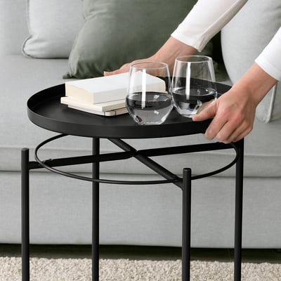 Person grabs a wine glass from a black GLADOM tray table with legs, holding a book and glasses next to a couch.