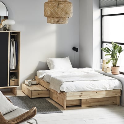 Minimalist bedroom with light wood GLAMBERGET extendable bed, white linens, grey rug, wicker pendant lamp, and potted plant near window.