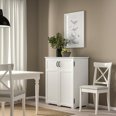 White GREÅKER cabinet with 4 doors, picture, and plant between window and dining table, with a chair beside.