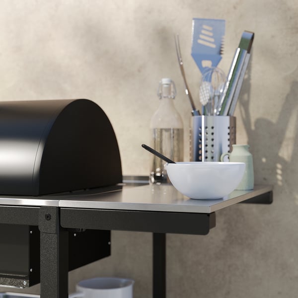 Modern black outdoor grill with stainless steel countertop, white bowl, utensils, against beige wall.