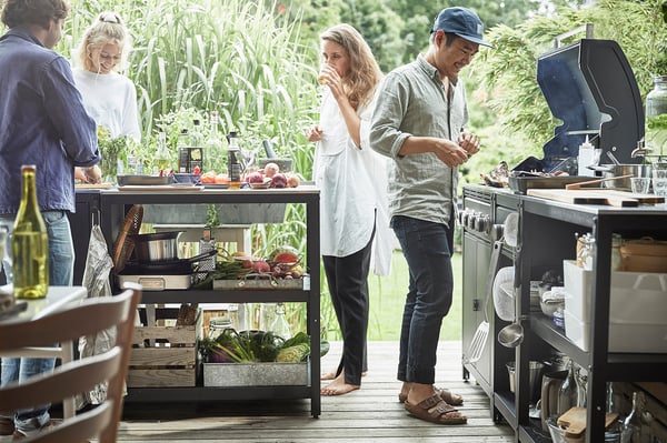 Four socialise around GRILLSKÄR steel kitchen island with veggies and grilling tools. Has galvanised shelves for outdoor cooking.