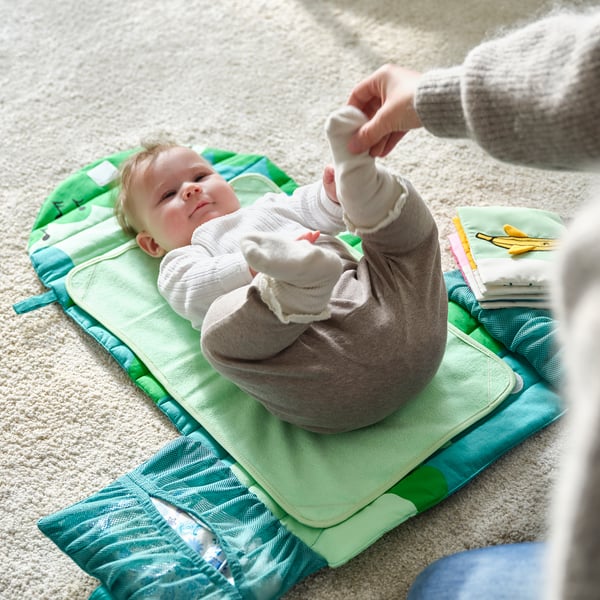 A baby lies on a green changing mat. An adults hand reaches towards the baby. The mat has pockets for diapers.