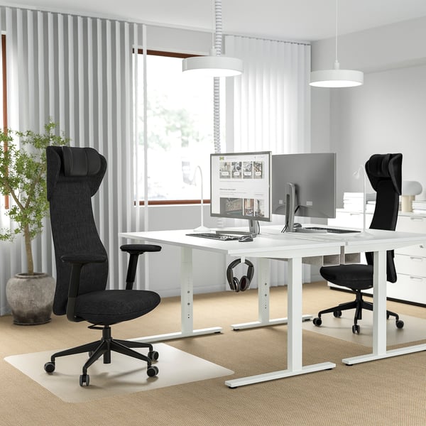 Modern office setup with two ergonomic black chairs at spacious desks with screens and tech. Bright room with large windows.