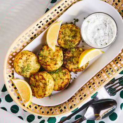 A plate of golden-brown vegetable cakes with lemon wedges and a side of white sauce. Served with silver cutlery.