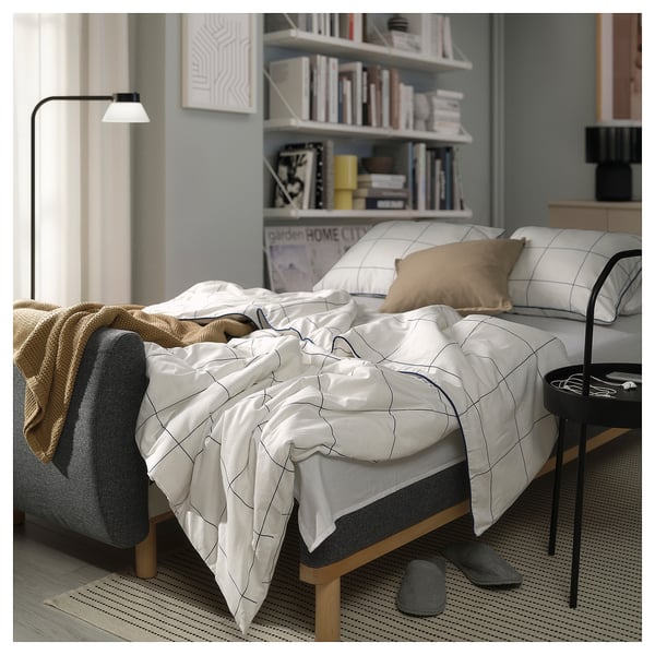 Modern bedroom with unmade GRUNNARP sofa-bed, white grid bedding, tan pillows, grey slippers, bookshelf, and floor lamp.