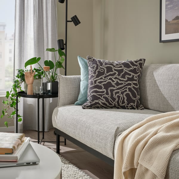 Cozy living room with GULDFLY cushion, gray couch, decor pillows, blanket, plants, laptop, books, framed picture, lamp.