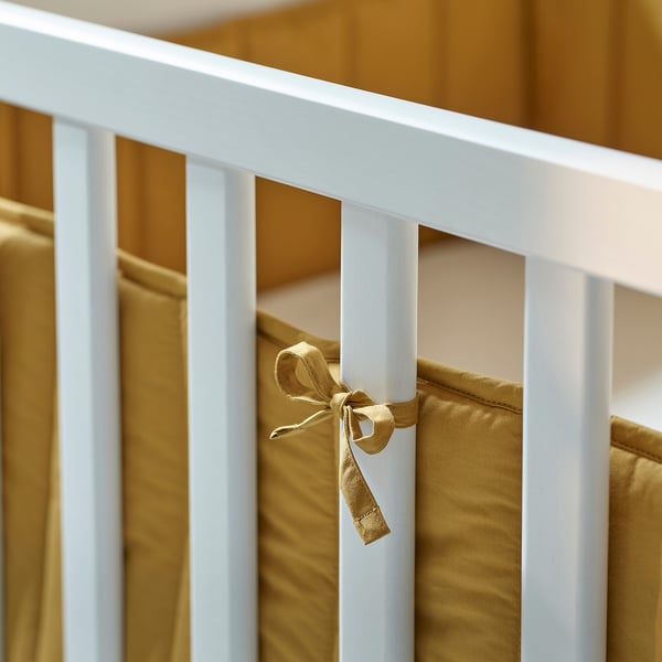 A white wooden crib with a gold-colored bumper pad tied to the slats, ensuring a soft barrier to prevent children from hitting the hard frame.