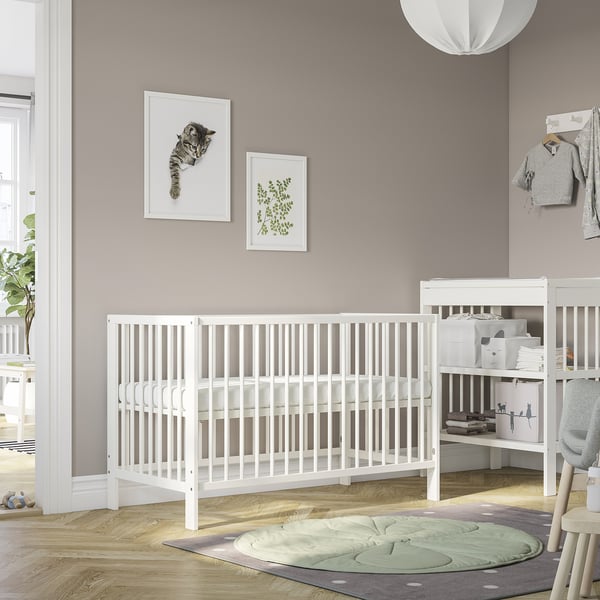 Baby room with white GULLIVER cot, grey walls, plant, art prints, and round rug. Cot is large, centered, with shelves beside it.