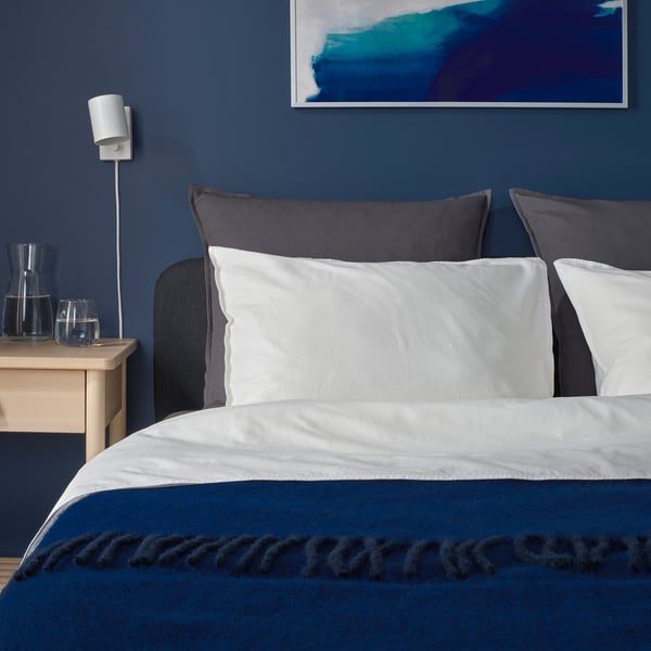 Modern bedroom with dark blue wall, GURLI grey cushions, white linens, wooden bedside table, blue glassware, wall lamp.