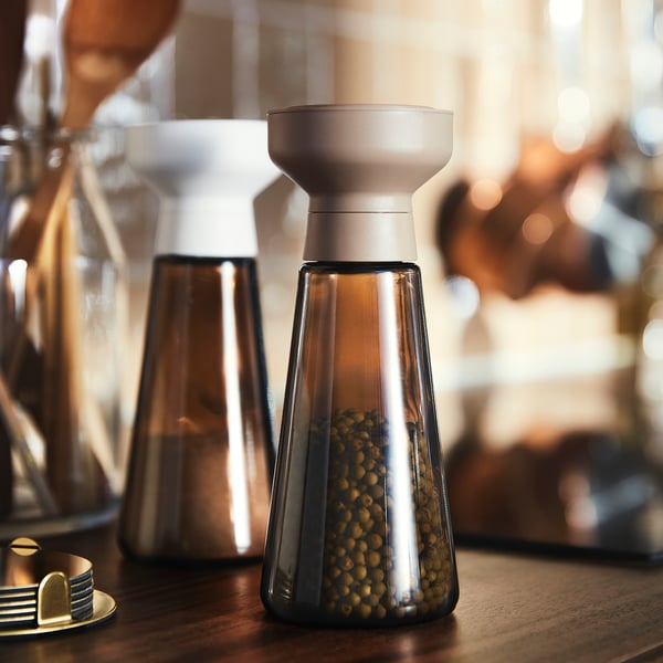 Two brown glass HALVTOM jars on wooden table, one filled with green peppercorns, topped with white grinders, designed for spice storage and grinding.
