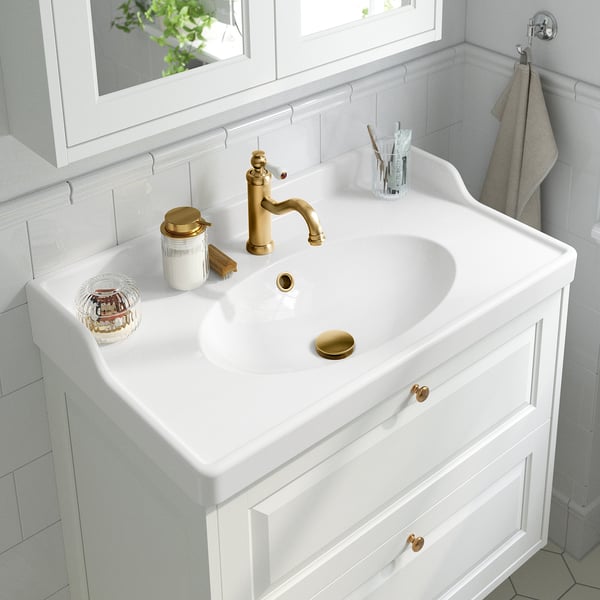 Elegant white bathroom vanity with gold-finished tap and drain. Functional items like toothbrush holder, soap dispenser, and towel.