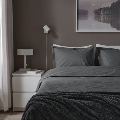 Bedroom with dark grey HAMPFLOCKEL duvet set. Modern white bedside lamp. Large framed art above bed. Curtain on left. Soft, textured blanket at foot.