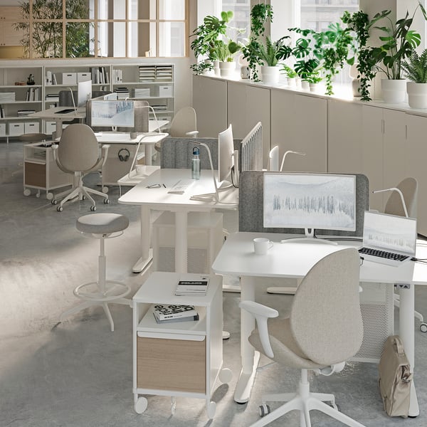Modern office space with white desks, ergonomic chairs, computers, and numerous potted plants. Clean, minimalist design with ample natural light.