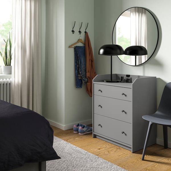 Modern bedroom with HAUGA grey chest, round mirror, black lamp, blue bed, wooden floor, and green walls.