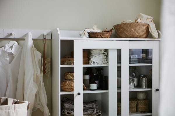 White HAUGA cabinet with glass doors displaying folded linens, jars, and baskets.