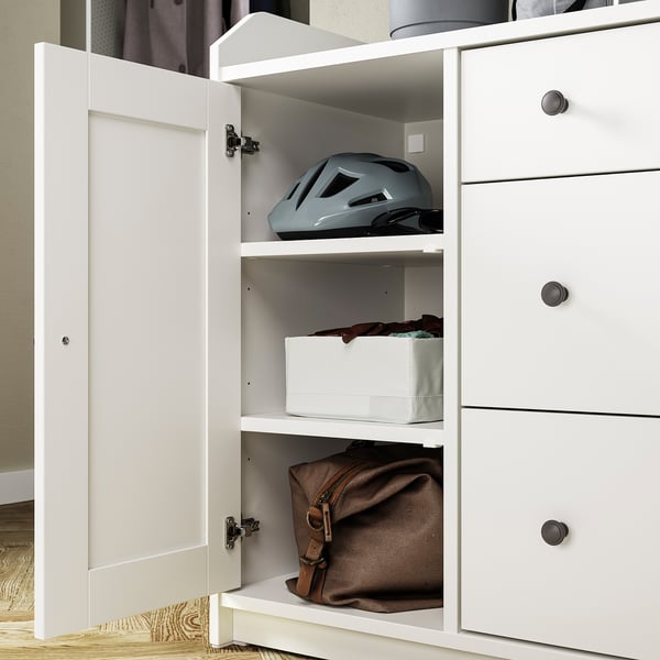 A white HAUGA sideboard with an open door revealing a shelf with a blue helmet and folded clothes above a brown bag.