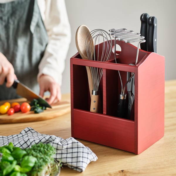 Red wooden kitchen utensil holder with knives, whisks, and spoons, on a wooden table with someone chopping vegetables nearby.