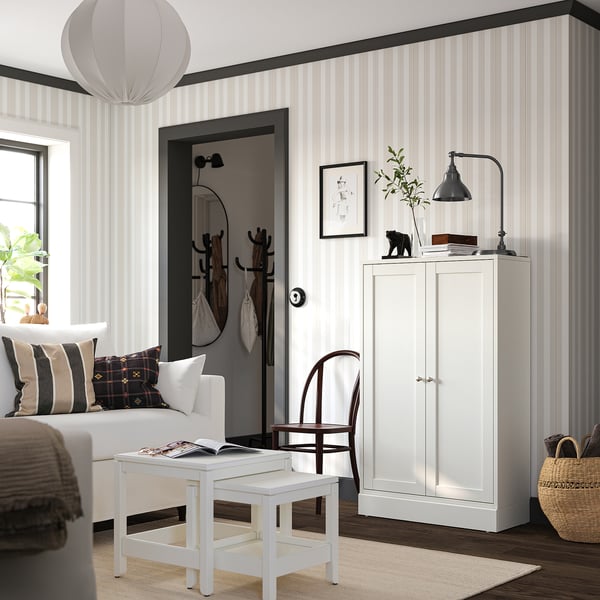 Elonged white storage cabinet with doors and shelves against striped wall, near a white couch with colourful pillows, lit by a black lamp.
