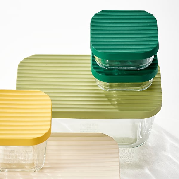 Three colourful, stackable glass containers with textured lids. One yellow, one green, and one transparent, arranged on a wooden surface.
