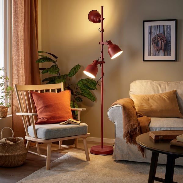 HEKTAR floor lamp in red stands tall in cosy living space, lighting a grey armchair and beige couch, complementing rustic decor.