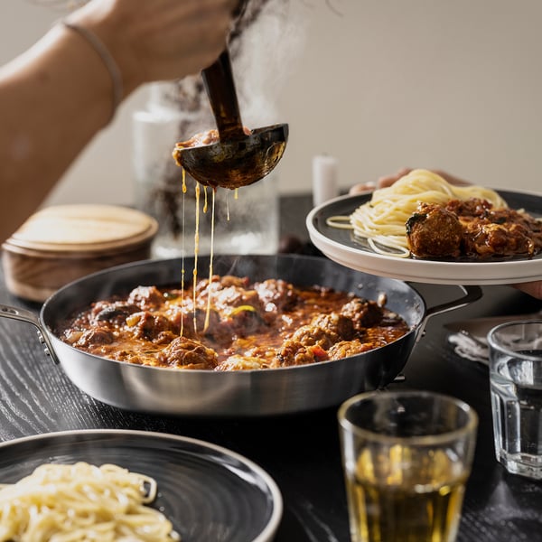 A person ladles a rich meat sauce from a stainless-steel pan, demonstrating its even heat distribution.