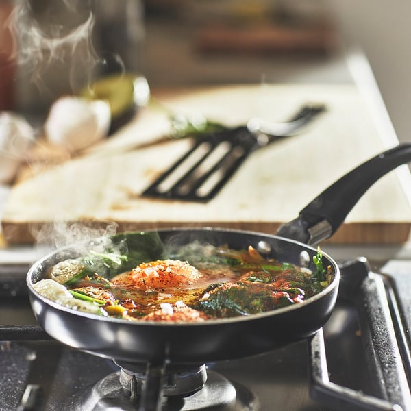 Black frying pan on stove, cooking spinach and tomatoes. White smoke rising, wooden cutting board with herbs behind.