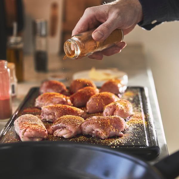 A hand seasoning raw chicken thighs on a baking tray with a non-stick ceramic coating.