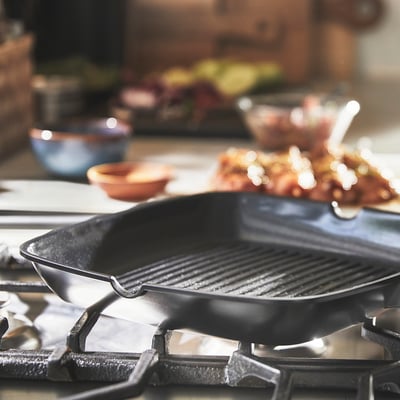 Grill pan with ribbed surface sits on gas stovetop, ready for cooking. Non-stick coating for easy food release and cleaning.